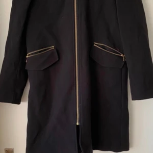 Svart kappa från H&M - En elegant svart kappa från H&M med dragkedja framtill och två fickor med dragkedjor. Jackan har långa ärmar och en rak passform.