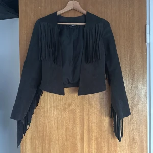 Svart mockajacka med fransar - En svart pu mockajacka med långa ärmar och fransdetaljer på både fram- och baksidan samt längs ärmarna. Jackan är kort och har en öppen design utan knappar eller dragkedja.