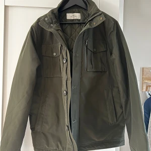 Grön jacka från John Henric - En grön jacka från John Henric i storlek M. Jackan har flera fickor, inklusive två bröstfickor med knappar och en dragkedjeficka. Den har en dragkedja och knappar för stängning samt en quiltad insida. fint skick använd fåtal gånger