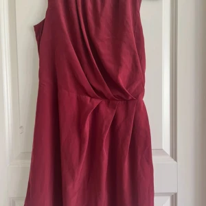 Vinröd klänning - En elegant vinröd klänning med draperad design. Klänningen har en ärmlös stil och en öppning i  ryggen.
