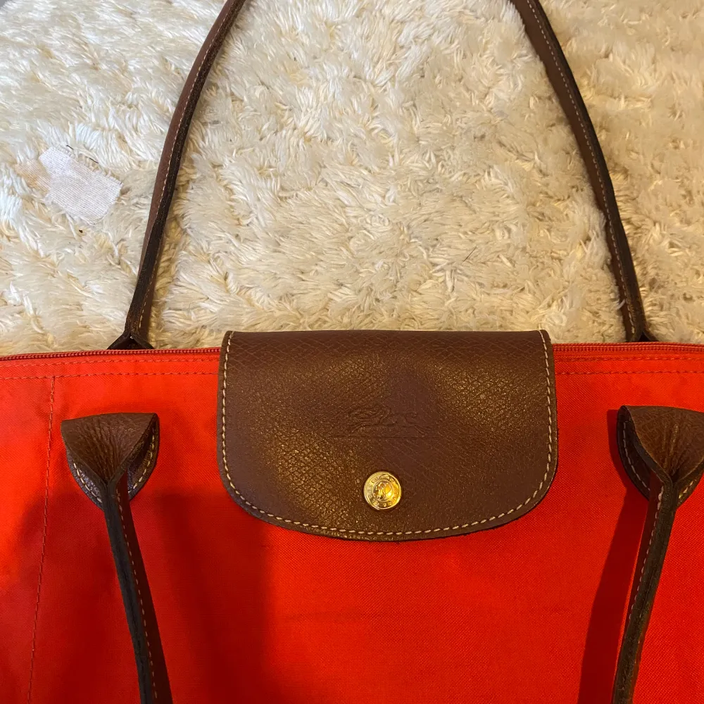 En snygg Loncchamp väska i knall orange färg. Modell Lapliage Väskan har tecken på användning där av priset. Läng:48 cm Bredd:32cm. Den har ett litet hål (se bild 5) och lite missfärgad. Hör av er vid frågor eller funderingar! 🧡🟠. Väskor.