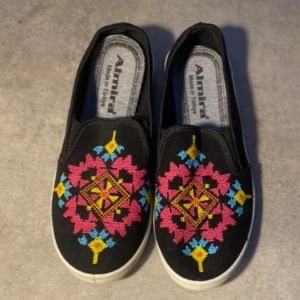 Slip on- svart  - Svarta slip on med broderi på framsidan av skorna. Skorna är i nytt skick och oanvända dock utan prislapp 