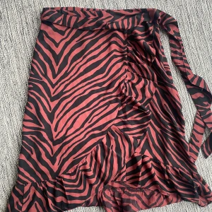 Svart och röd omlott kjol i leopardmönster i strl xs - Svart och röd omlott kjol i leopardmönster i strl xs.  Förutom en liten tråd se bild 2 är kjolen i fint begagnat skick då den endast använts en gång.  Mått Midjan: ca 32 cm Total längd: ca 60 cm