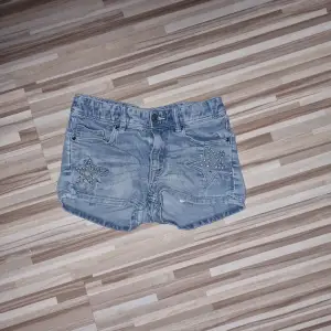 Ett par blåa jeansshorts med stjärndetaljer på fram- och baksidan. Shortsen har en knapp och dragkedja framtill samt fickor både fram och bak. Från H&M i storlek 134.