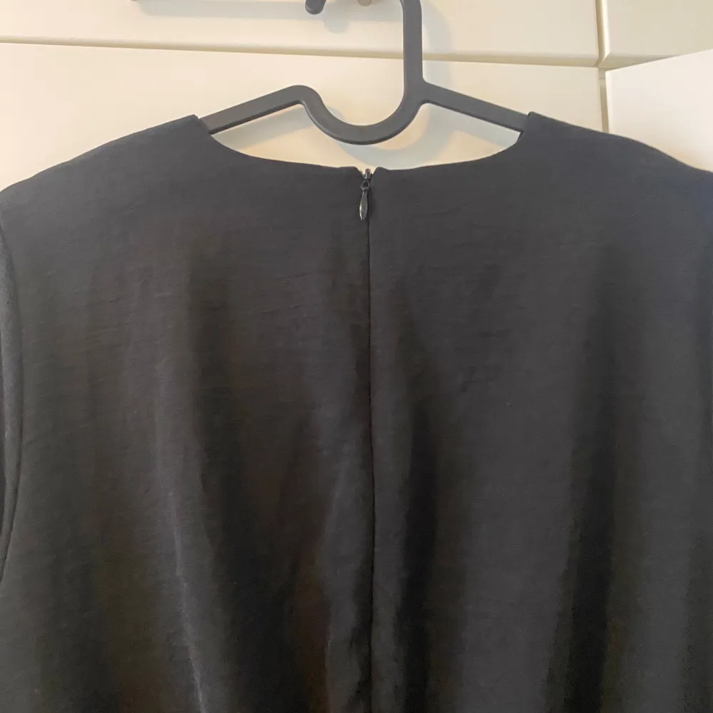 En svart slät blus med en dragkedja bakom ryggen och små detaljer från bröstet till midjan. Den är mellan lång blus som sitter vid midjan och som är löst nedåt. Puserot.