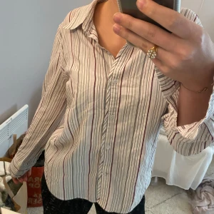 Randig vit skjorta - Här säljer jag min randiga vita skjorta. Den står att den är i storlek XL vilket jag har svårt att tro på. Mig passar den perfekt och jag storlek S/M vanligtvis. Den har vinröda och gråa ränder på sig och kommer från märket Emilia.