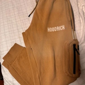 Bruna mjukisbyxor från Hoodrich - Ett par bekväma bruna mjukisbyxor från Hoodrich med elastisk midja och dragsko. Byxorna har en ficka med dragkedja på sidan och en vit logotyp på vänster ben.