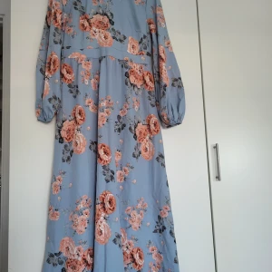 Blommig klänning  - En vacker klänning med blommigt mönster i blått och rosa. Klänningen har långa ärmar och en rund halsringning med en volang. Den har en lös passform och är perfekt för både vardag och fest. Används en gång bara!