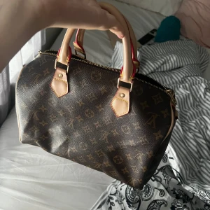 Brun handväska med monogram - En elegant brun handväska med monogrammönster och ljusa handtag. Väskan har en dragkedja och detaljer i ljusare färg.