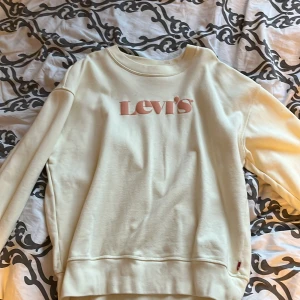 Vit sweatshirt från Levi's - En vit sweatshirt från Levi's med rosa logotyp på framsidan. Tröjan har långa ärmar och ribbstickade muddar vid ärmslut och nederkant.