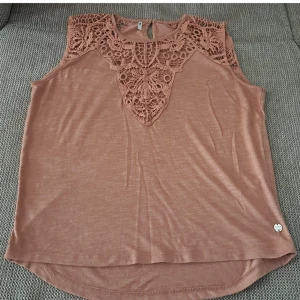 Brun ärmlös topp med spetsdetaljer - En brun ärmlös topp med vackra spetsdetaljer runt halsen och axlarna. Toppen har en rund halsringning och en knapp i nacken för enkel på- och avtagning. Materialet är mjukt och bekvämt, perfekt för både vardag och fest.