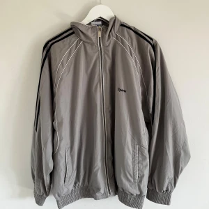 Vintage Streetwear Vindjacka - En grå supercool vintage streetwear vindjacka från SportsWear med dragkedja framtill. Jackan har långa ärmar och ribbstickade muddar vid ärmslut och nederkant. Den har också två fickor framtill och texten 'Sports' broderad på bröstet och ryggen.