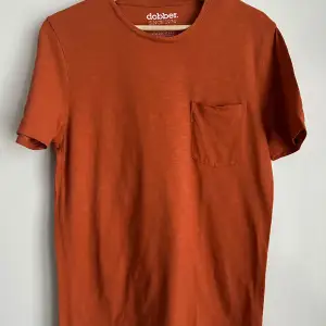 En enkel och stilren orange t-shirt från Dobber med en bröstficka. T-shirten har korta ärmar och rund halsringning.