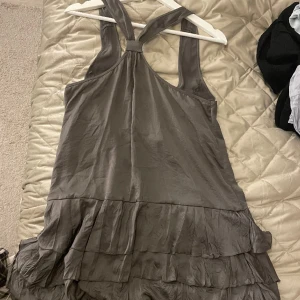 Grön/grå klänning med volanger - Grön klänning i glansigt material med volanger, dragkedja och korsad hals. Oklart vilken storlek men har själv s/m. 