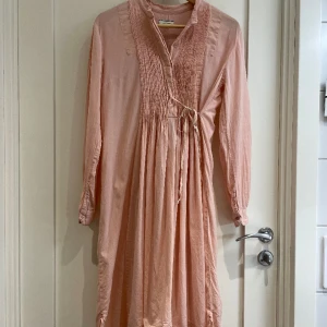 Underbar By Malene Birger-klänning i stl 36 - Skir, rosa klänning, lite längre baktill än fram, med smock och knappar över bröstet. By Malene Birger. Superfin året om beroende på hur du stylar den.  Knappt använd, inga anmärkningar finns, absolut i nyskick. Stl 36. 