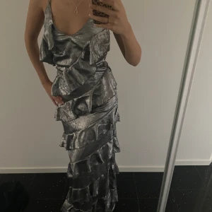 Silvrig balklänning  - Säljer denna asfina silvriga balklänning med volanger. Har en liten slits på ena sidan vilket gör det enklare att gå. Justerbara band. Storlek 38 och sitter bra på mig som har S!