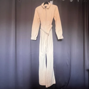 Beige jumpsuit från BikBok - En beige jumpsuit från BikBok i storlek S. Jumpsuiten har långa ärmar och en dragkedja framtill. Den är tillverkad i ett ribbat material och har ett bälte i midjan för en justerbar passform.