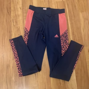 Blå och rosa leggings från Adidas - Ett par blå leggings från Adidas med rosa detaljer och leopardmönster längs sidorna. Leggingsen har en elastisk midja och är designade för en bekväm passform. Säljs för att de blivit för små💕