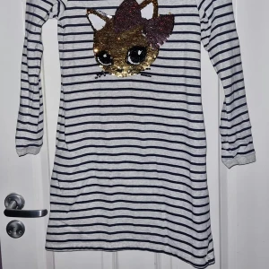 Randig klänning med paljettmotiv - En grå och svart randig klänning med långa ärmar och ett stort paljettmotiv (vändbar) av en katt på framsidan. Klänningen har en rund halsringning och är tillverkad i ett mjukt och bekvämt material. Från H&M i stl 146/152