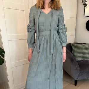 Bridgerton inspirerad klänning - Turkos midiklänning inspirerad av Bridgerton. Vad/ankellång på personen, 169cm. Snören vid halsen går att ha både hängande eller knutna i en rosett. Volang på ärmarna samt små rosetter. Trekvartslånga ärmar.