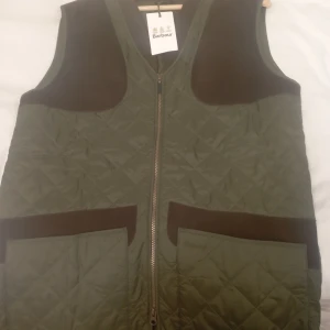 BARBOUR KEEPERWEAR GILET - (Oanvänd ny ) Traditionell väst i countrystil från Barbour. Utformad för att passa skytten, men fungerar lika bra för dig som söker en snygg väst att använda till vardags. Quiltad och lätt fodrad väst med dragkedja, känd som känd som "GW-västen" .
