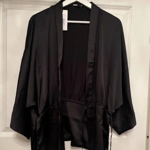 Omlottblus - Svart omlottblus med vida ärmar från BikBok i storlek XS men passar även större. Aldrig använd, prislapp finns kvar. Nypris 349 kr. Stängs med två knytningar, varav en som syns.  Pris kan diskuteras. Hör gärna av er för fler bilder eller frågor. 