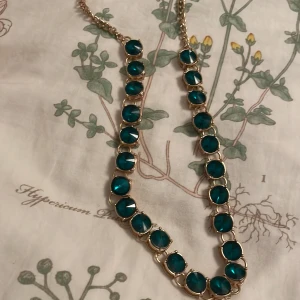 Guldhalsband med gröna och klara stenar - Säljer ett elegant halsband med gröna och klara stenar. Halsbandet har en kedja som är guldfärgat och är dekorerat med runda gröna stenar på ena sidan och klara stenar på andra sidan. Perfekt för att lägga till en touch av färg och elegans till din outfit. Köp direkt eller ge prisförslag 💛 är osäker på varumärket