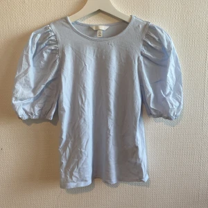 Ljusblå t-shirt med puffärmar  - Säljer en söt ljusblå topp med puffärmar. Toppen är i storlek XS och är perfekt för både vardag och fest. Den har en rund halsringning och är tillverkad i ett mjukt och bekvämt material. Passar bra till både jeans och kjol!