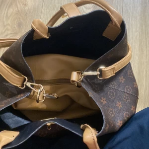 Brun och beige axelväska med monogram - Säljer en snygg brun och beige axelväska med monogrammönster. Väskan har två långa handtag och en dragkedja på insidan. Den är rymlig och perfekt för vardagsbruk. Väskan är i bra skick och har en stilren design med gulddetaljer.