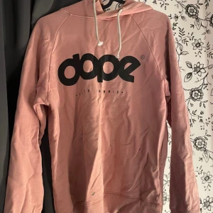 Rosa hoodie från Dope - Säljer en snygg rosa hoodie från Dope. Den är i storlek xxs och har en stor svart logga på bröstet. Hoodien har långa ärmar och en justerbar huva med vita snören. Perfekt för en avslappnad och cool stil! Finns dock en liten fläck längst ner på tröjan.