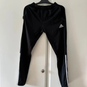 Svarta träningsbyxor från Adidas - Svarta träningsbyxor från Adidas med vita ränder längs sidorna och en dragkedja vid bensluten. Byxorna har en elastisk midja och Adidas-logotypen på vänster ben.