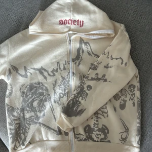 Beige jacka med skelettmönster från Society - En beige hoodie med dragkedja från Society. Jackan har ett unikt skelettmönster i grått och texten 'Society' i rött på huvan. Den har långa ärmar och ribbstickade muddar vid ärmslut och nederkant. vet inte om den är äkta eller inte 💕står ingen storlek men sitter som L