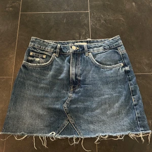 Blå jeanskjol från Trafaluc - Säljer en snygg blå jeanskjol från Trafaluc. Kjolen är i storlek S och har en rå kant nertill som ger en cool och avslappnad look. Den har fem fickor, två fram och två bak, samt en liten myntficka fram. Kjolen har en knapp och dragkedja framtill. Perfekt för både vardag och fest!