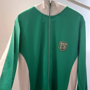 Marwin Sports kofta - Säljer en grön och vit vintage kofta från Marwin Sports. Jackan har en dragkedja framtill och ett emblem på bröstet med texten 'M.W.N'. På baksidan finns en stor text med 'Marwin'. Perfekt för sportiga aktiviteter eller som en snygg vardagskofta💚