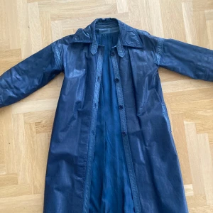 Blå skinnkappa - Lång fodrad skinnkappa i äkta läder. Mörkblå med ljusare blå detaljer längs med knappar, krage och ärmar. Ser ut såhär på mig (158 cm, storlek S) men passar storlek M