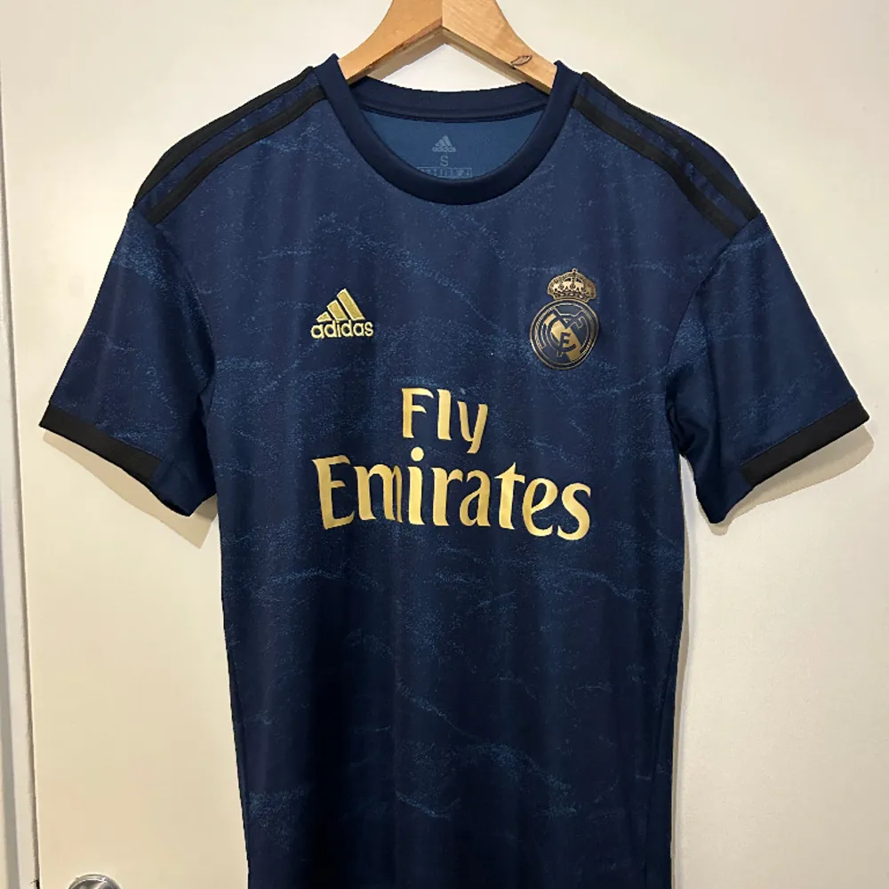 Äkta mörkblå Real Madrid 2019/20 borta fotbollströja med Hazard och nummer 7 på ryggen. Guldig text och detaljer samt svarta ränder på ärmarna i storlek S. Använd få gånger. . T-shirts.