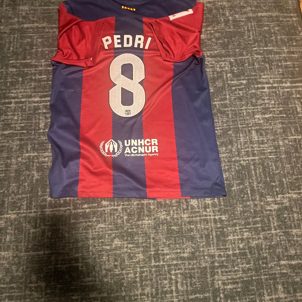 Tjena, säljer min Barcelona tröja med Pedri 8. P:et i Pedri är tejpat och strykt med strykjärn så att det sitter fast pga av att det förra lossnade…. T-shirts.