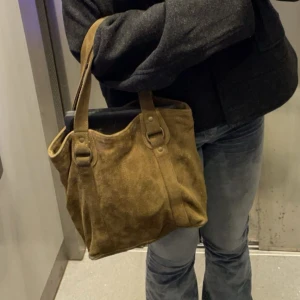 Brun mocka axelväska - En brun axelväska i mocka med två handtag. Väskan har en rymlig design och är i gott skick.