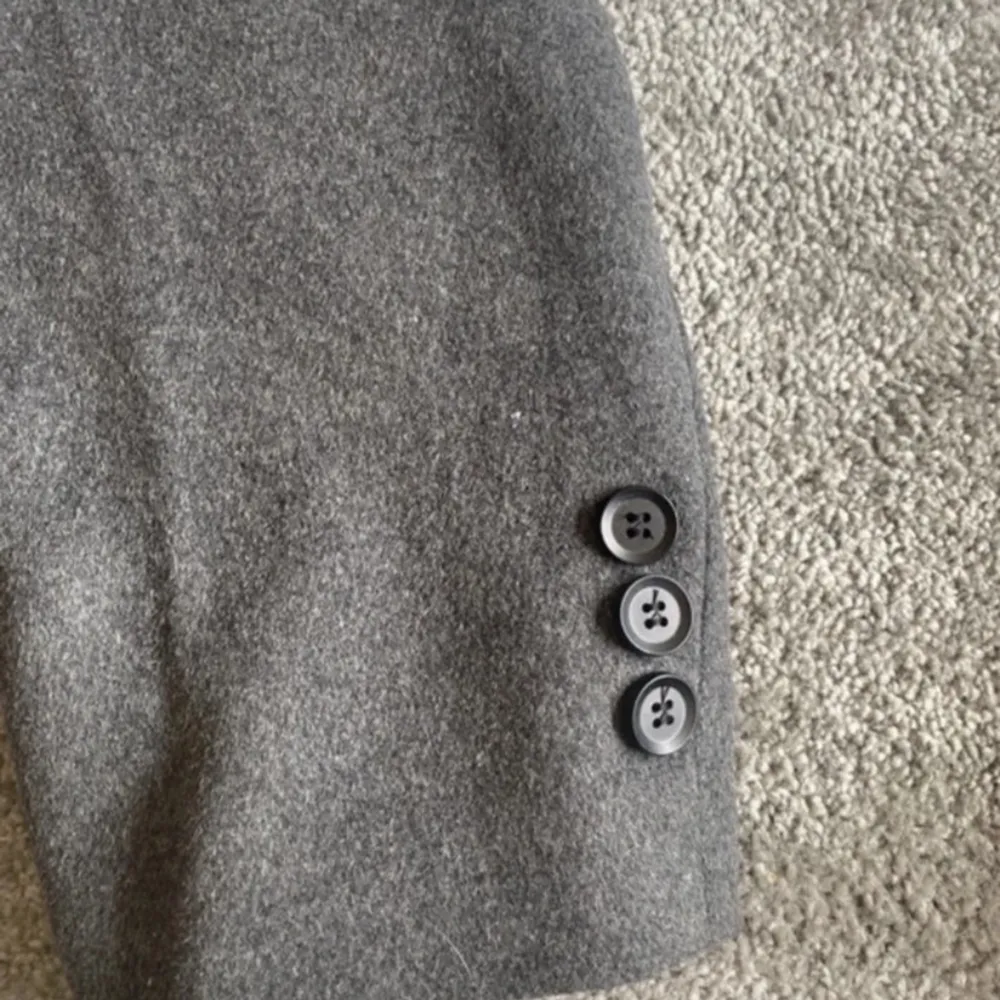 En stilren grå rock med knappar framtill och på ärmsluten. Rocken har en klassisk design med en krage och är tillverkad i ett mjukt material. En knapp (se bild) har gått sönder. Annars är kappan endast använd 1 gång . Takit.