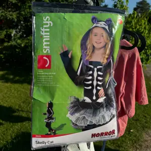 En svart och vit Skelly Cat kostym från Smiffys, inkluderar en klänning och en bolero med huva. Kostymen har ett skelettmönster och detaljer i lila och vitt. Perfekt för Halloween eller andra temafester.