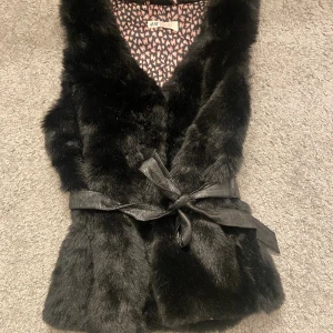 Fluffig svart väst  - Svart fluffig väst man kan knyta den och inuti är det rosa hjärtan men man ser dom inte när man har på den.Det är storlek 11 till 12 men jag tycker att det kan vara 9 till 13.Från H&M. 