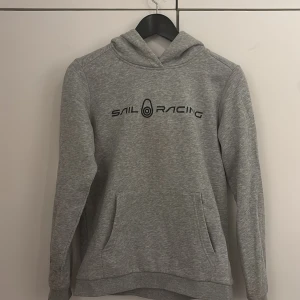 Grå hoodie från Sail Racing - Säljer en snygg grå hoodie från Sail Racing i mycket bra skick. Den har en klassisk design med en stor ficka fram och logga på bröstet. Perfekt för både vardag och chill. Passar bra till höst och vår!