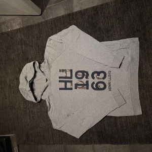 Grå hoodie från Henri Lloyd - Säljer en snygg grå hoodie från Henri Lloyd i storlek 14-15/170.Den har ett coolt tryck på framsidan med texten 'HL 1963'. Perfekt för en avslappnad stil och passar bra till både jeans och joggers. Huvan är mjuk och bekväm, perfekt för kyliga dagar.