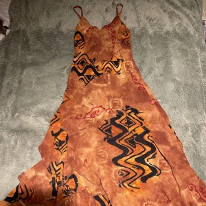 Brun mönstrad klänning med asymetriska volanger - Säljer en unik brun vintage klänning med ett abstrakt mönster i svart och orange från 90talet. Klänningen har smala axelband och asymmetrisk volangdetaljer i flera lager. Perfekt för sommarens fester eller en bohemisk look. Materialet känns lätt och luftigt.