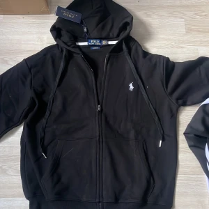 Svart hoodie från Polo Ralph Lauren - Säljer en snygg svart hoodie från Polo Ralph Lauren i storlek S. Den har en dragkedja framtill och en klassisk logga på bröstet. Perfekt för både vardag och chill. Huvan har dragsko och det finns fickor framtill. Passar perfekt till höst och vår!