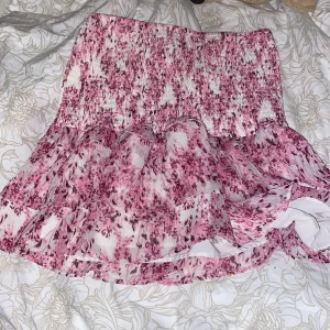 Rosa blommig kjol med volang - Superfin rosa blommig kjol med volangdetaljer. Perfekt för sommardagar eller en festlig kväll. Den har en elastisk midja för bekväm passform och är gjord i ett lätt och luftigt material. Kjolen är kort och har ett romantiskt mönster som verkligen sticker ut. ❤️