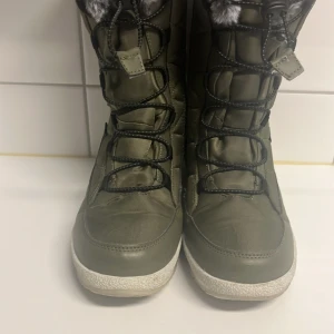 Olivgröna vinterstövlar med fuskpäls - Säljer ett par olivgröna vinterstövlar i mycket bra skick. De har snörning och är fodrade med grå fuskpäls för extra värme. Perfekta för kalla vinterdagar och har en robust sula för bra grepp. Stövlarna är både snygga och praktiska för vintern!
