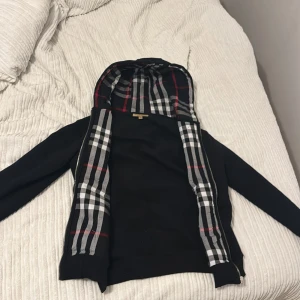 Svart jacka från Burberry - Säljer en snygg svart kofta från Burberry med klassiskt rutigt mönster i fodret. Koftan har långa ärmar och dragkedja framtill. Perfekt för höst och vår! 🖤