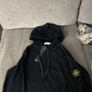 Säljer en snygg svart hoodie från Stone Island. Den har en klassisk design med en känguruficka och justerbar huva. Perfekt för både vardag och chill. Märkets ikoniska logga sitter på ärmen. Passar perfekt till höst och vinter!