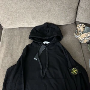 Svart hoodie från Stone Island - Säljer en snygg svart hoodie från Stone Island. Den har en klassisk design med en känguruficka och justerbar huva. Perfekt för både vardag och chill. Märkets ikoniska logga sitter på ärmen. Passar perfekt till höst och vinter!